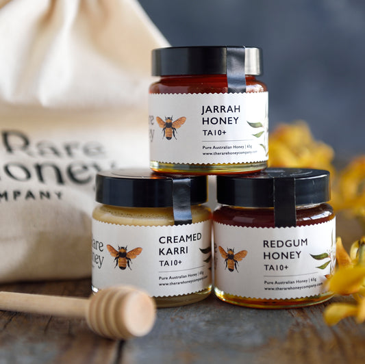A set of three jars of honey, each labeled differently, arranged in front of a reusable gift bag with the brand name 'Rare Honey'. The honey jars are placed on a wooden surface with a honey dipper and yellow flowers in the background.