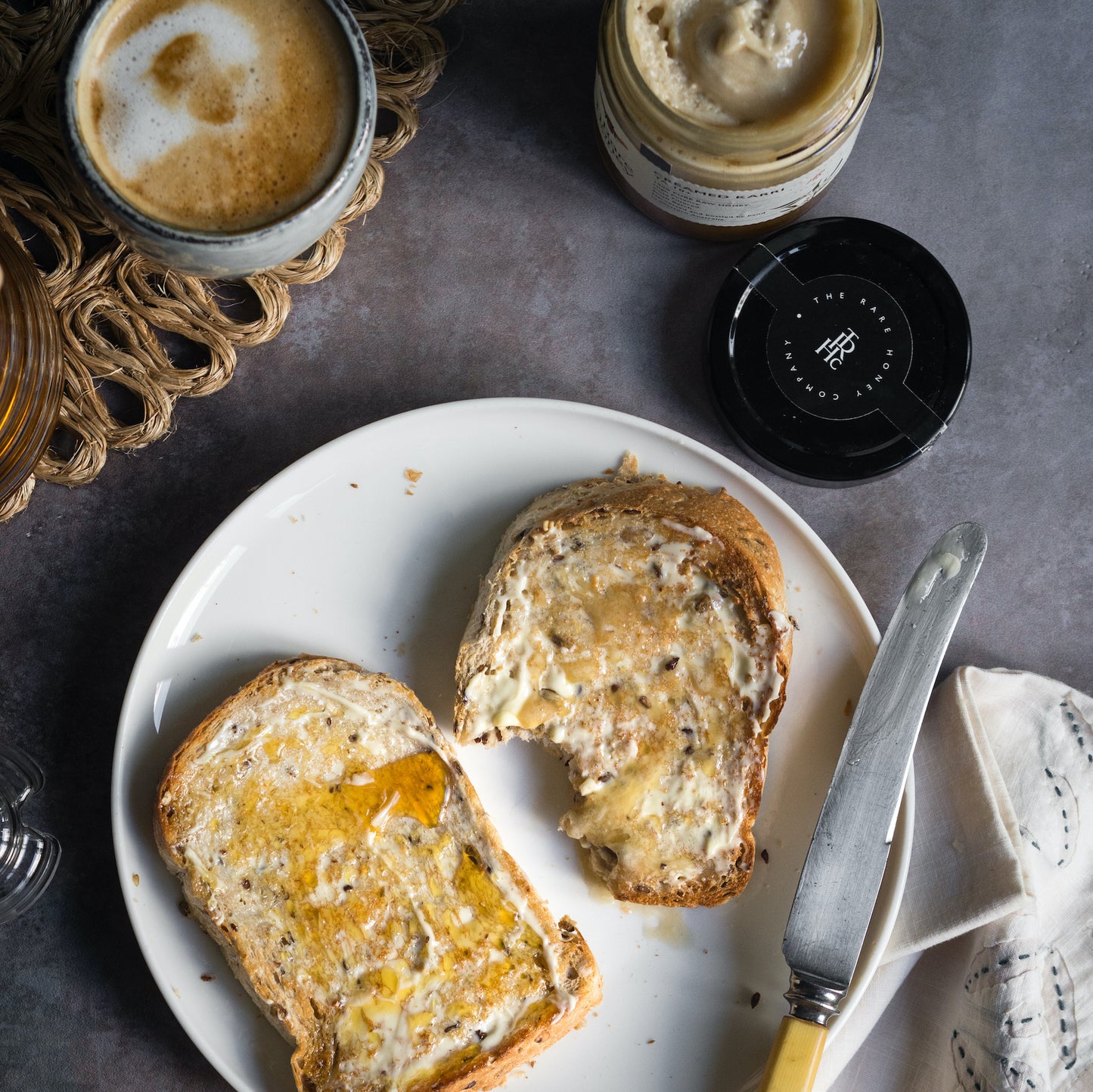 Premium Australian honey on toast with butter placed on a white plate with a cup of coffee in the top left corner and an open jar of The Rare Honey Creamed Karri honey in right.