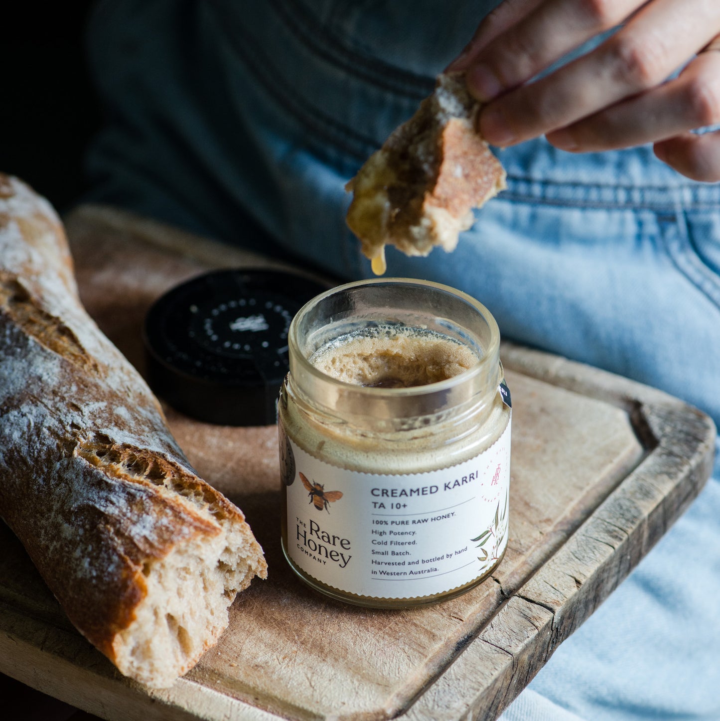 Person spreading honey on bread with a jar of The Rare Honey Company creamed karri honey on a wooden board.